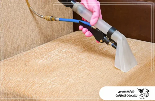 تنظيف مجالس قماش بالدمام 0557756533 شركة الفارس لقطة مقربة ليد مرتدية قفازًا ورديًا وهي تستخدم أداة استخلاص متخصصة لتنظيف مقعد كنبة بلون بيج فاتح.