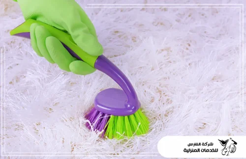 أرخص شركة تنظيف بالدمام 0557756533 شركة الفارس يد ترتدي قفازاً أخضر تفرك سجادة بيضاء ناعمة بفرشاة ملونة.