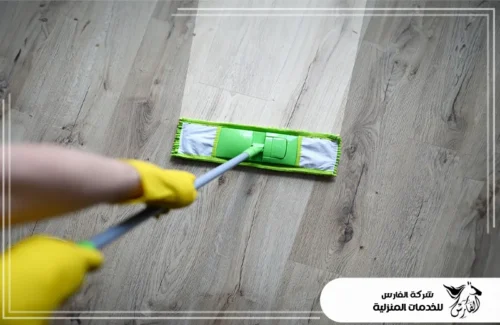 تعقيم المنازل 0557756533 شركة الفارس لقطة علوية تظهر مسح الأرضيات الخشبية بممسحة عصرية، مع قفازات صفراء للحماية.