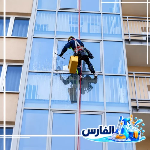 تنظيف نوافذ المباني الخارجية بالرياض باستخدام الحبال عامل ينظف النوافذ الزجاجية الخارجية لمبنى متعدد الطوابق باستخدام الحبال وأدوات التنظيف، مع ظهور شعار شركة الفارس.